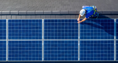 A worker in safety gear installs or inspects blue solar panels on a sloped gray tiled roof.