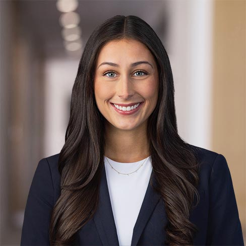 A woman with long brown hair wearing a navy blazer and white top smiles in a hallway with blurred lights in the background.