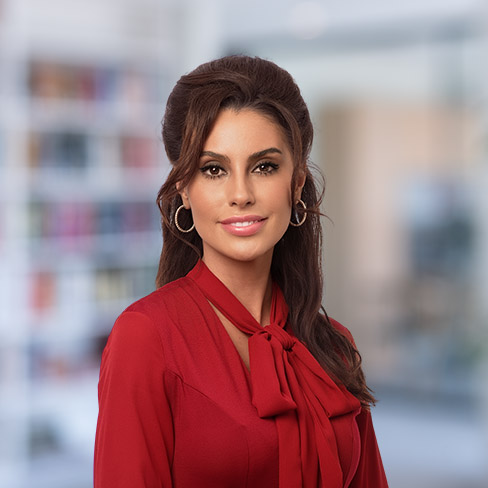 A woman with long brown hair, wearing a red blouse with a bow and hoop earrings, stands in a blurred office or library setting.